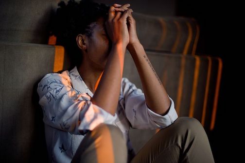 A young person sitting in a closed position and holding their own hands in front of their face, expressing emotional pain. 