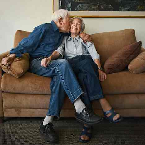 Elderly couple embracing one another on a couch, the man is kissing the woman and she is joyful.  