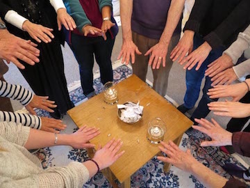 A group of 9 people standing in a circle and putting their hands towards a table, the table has two lit candles and a pile of small pieces of paper in the middle. 