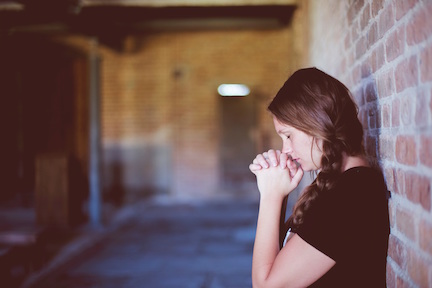 Woman standing with her back against a wall, clasping her hands in prayer. 