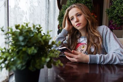 Young woman sitting at a table and looking sad while holding her cell phone. 