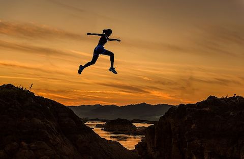 An athletic woman jumping from one cliff to another with a sunset in the background. 