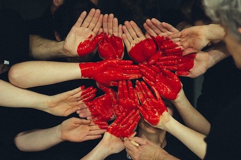 A small group of people putting their hands together to illustrate a heart made out of red paint on their hands. 