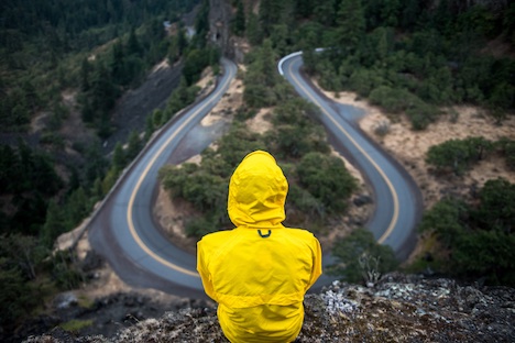 Rear view of a person in a yellow rain jacket looking down at a looping mountain road. 