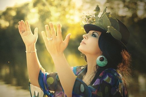 Woman wearing a purple and blue shaw and a bowler hat with leaves on it, looking upward with open hands. 