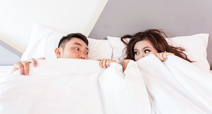 A couple looking at each other while in bed, covering their faces with the blankets. 