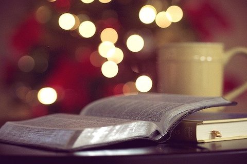 One opened book next to one closed book sitting on a desk with lights in the background. 