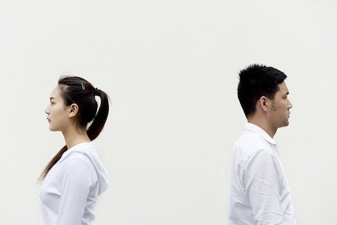 A man and a woman facing back to back wearing white clothes in front a white wall. 