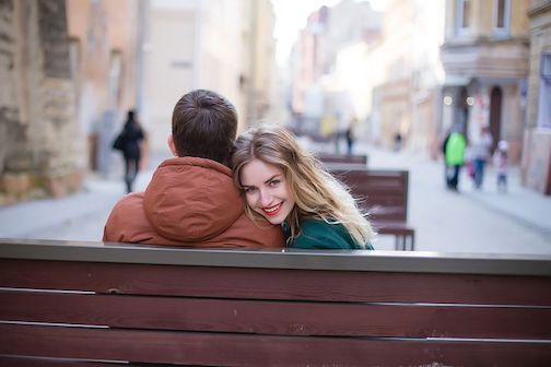 A couple sitting on a bench in a city, the woman is looking at the camera and smiling. 