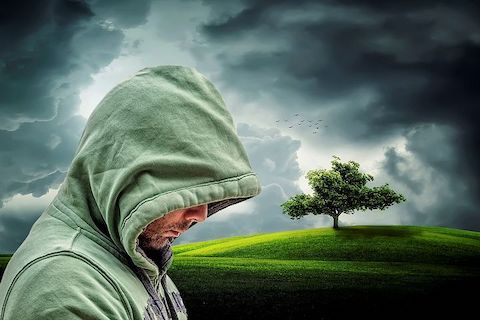 Hooded man looking downward at the floor, with a stormy sky and a tree in the distance. 