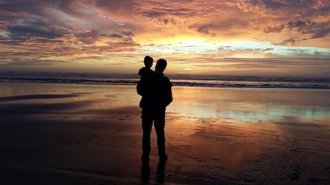 A silhouette of a man holding his toddler child in his arms on a beach viewing a sunset. 