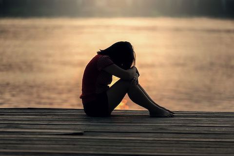 A woman sitting down on a dock and sulking with her head down. The background is a sunset and calm water in the background. 