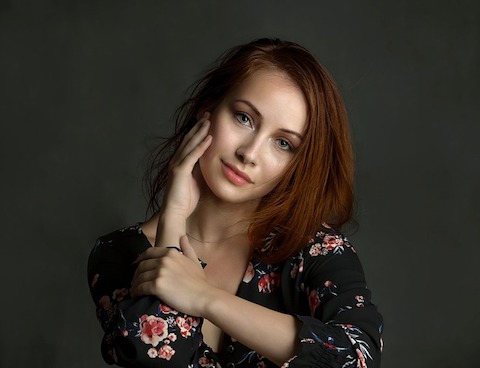 Red haired woman wearing a black shirt with flowers on it,  gently posing with her hand on her face. 