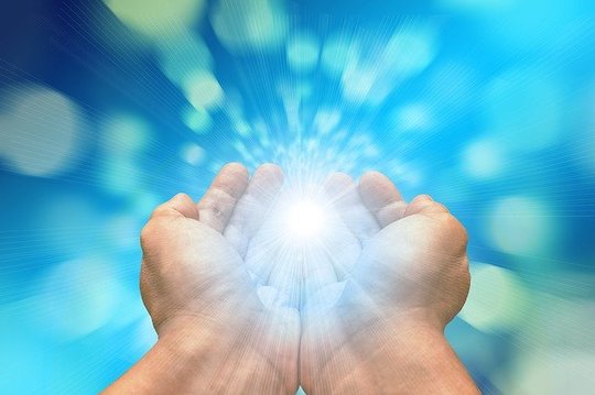 Two adjoined hands exuding light from center of palms in front of a bright blue background. 