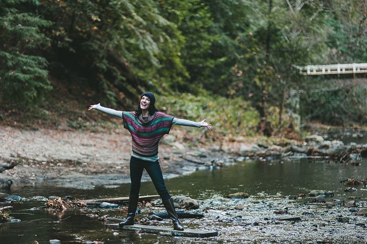 A woman on a riverbank smiling and stretching her arms out to the side. 