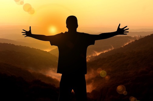 A silhouette of a man holding his hands outward and facing a sunset. 