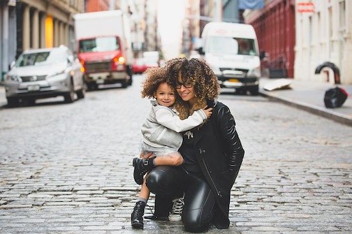 Woman holding her toddler and smiling into the camera, kneeling in the middle of a downtown street. 