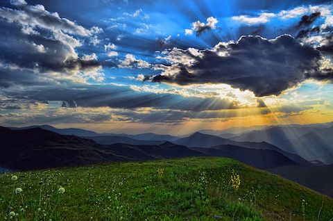 A view of the horizon, the sun is shining behind a daytime sky over a mountain range from the viewpoint of a grassy hill. 