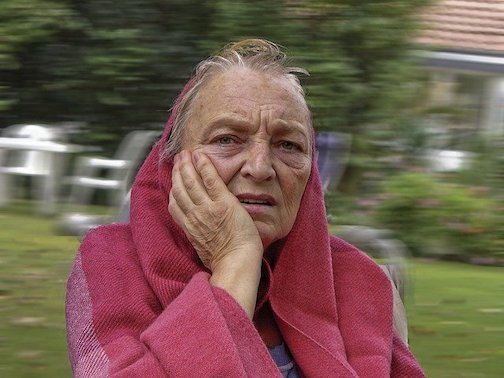Elderly woman wearing a red hooded sweatshirt, holding her face with one hand. 