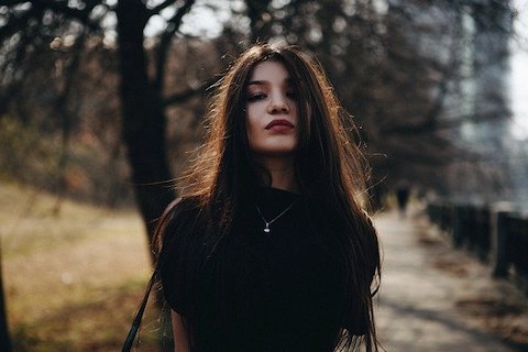 Woman with long hair wearing dark colors, posing with her hands behind her back and staring into the camera. 