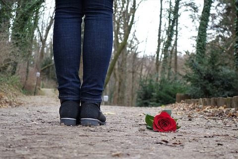 rear view of a person standing on a dirt trail in the woods with a rose on the ground. 