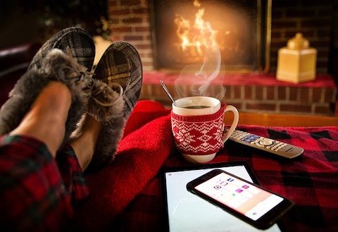 Close up of someone crossing their feet on a table in front of a fireplace. On table is a cup of hot chocolate, a remote control, and a smartphone. 