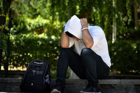 Man sitting on a curb next to his backpack. His hood is over his head and he is looking at the floor. 