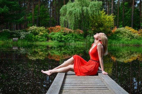 Woman wearing a reddish orange dress relaxing on the dock of a pond in a wooded area. 